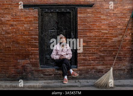 Dunbar Square, Bhaktapur, Nepal - 11 febbraio 2025 - Un uomo nepalese che riposa sulla strada sedeva su un passo accanto a un'antica porta Foto Stock