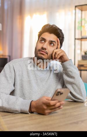 Giovane indiano seduto sul divano a tavola, scrivendo attivamente sullo smartphone, appoggiando la testa a mano nel soggiorno di casa. Il ragazzo arabo sembra concentrato, profondamente impegnato nella conversazione o nel compito Foto Stock