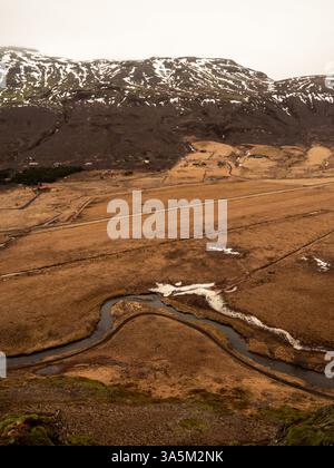 Una vastissima valle islandese con un fiume serpeggiante e aspre montagne con macchie di neve. Il paesaggio marrone-dorato contrasta con il vulcano scuro sl Foto Stock