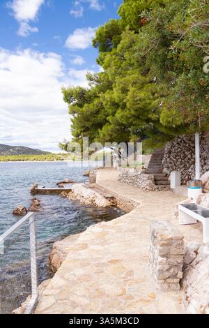 Tribunj, Croazia - 14 settembre 2024: Passerella costiera pavimentata di fronte ai giardini di fronte alle case con vegetazione Foto Stock