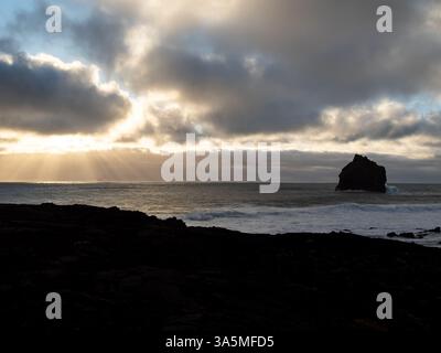 Una pila di mare solitaria si erge contro cieli spettacolari e raggi di sole dorati che penetrano attraverso le nuvole a Valahnúkamöl, la costa frastagliata dell'Islanda Foto Stock