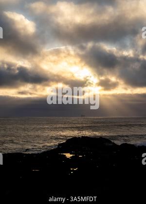 I raggi d'oro attraversano le nuvole spettacolari sull'Atlantico settentrionale a Valahnúkamöl, Islanda. Un paesaggio marino tranquillo con rocce lontane e riflessi Foto Stock