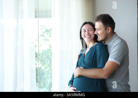 Felice donna asiatica incinta che guarda il suo partner in piedi accanto a una finestra a casa Foto Stock