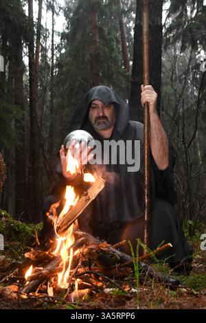 Un monaco dalle vesti nere siede accanto a un fuoco nella foresta e guarda in una palla di divinazione. Misticismo e magia, stregoni e streghe 3 Foto Stock