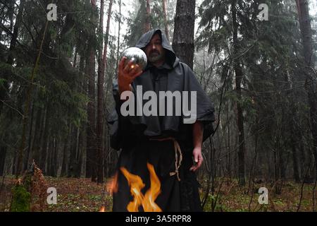 Un monaco dalle vesti nere siede accanto a un fuoco nella foresta e guarda in una palla di divinazione. Misticismo e magia, stregoni e streghe. Foto Stock