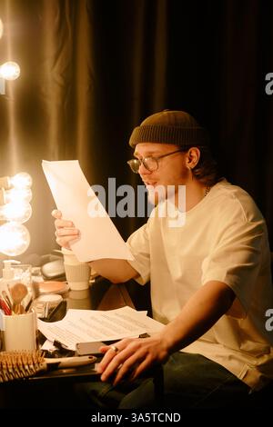 Un giovane seduto nello spogliatoio che legge un documento sotto le luminose luci del trucco, concentrato sui dettagli, circondato da cosmetici e pennelli, indossa un abbigliamento casual Foto Stock