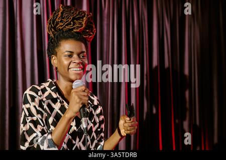 Donna afroamericana sorridente e tiene in mano il microfono mentre è sul palco con sfondo rosso, mostrando energia positiva nelle prestazioni Foto Stock