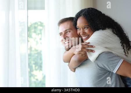 Divertente coppia interrazziale che si diverte a guardare attraverso una finestra scherzando a casa Foto Stock