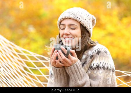 Donna in autunno in una foresta che beve un caffè seduto su un'amaca Foto Stock