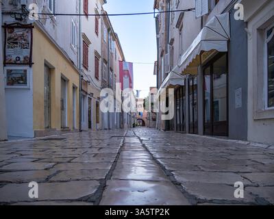 Rab, Croazia - 10 agosto 2024: Una stretta strada acciottolata costeggiata da edifici storici, con negozi e una bandiera appesa sul vicolo. T Foto Stock