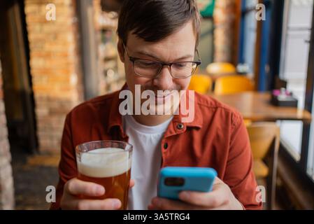 Uomo felice che si gode una pausa in un pub, sorseggiando una birra fredda mentre usa il suo smartphone. Sorridente e rilassato, sfoglia i social media sul suo dispositivo mobile, abbracciando il tempo libero Foto Stock