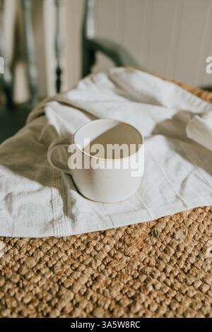 Una tazza di caffè bianca è posta su un asciugamano bianco su un tappeto marrone. La tazza è vuota e l'asciugamano è ben piegato. La scena offre un ambiente accogliente e rilassato Foto Stock