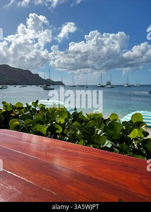 Soleggiata costa tropicale con barche a vela in una baia tranquilla, vista da un tavolo di legno vicino agli ombrelloni e alla vegetazione lussureggiante. Foto Stock