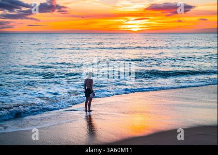 Un uomo cammina lungo la riva del mare in un bellissimo tramonto arancione sul mare Foto Stock