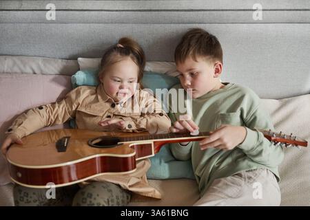 Ritratto ad alto angolo di una ragazza carina con sindrome di Down che suona la chitarra con il fratello mentre si sdraia a letto a casa, copia spazio Foto Stock