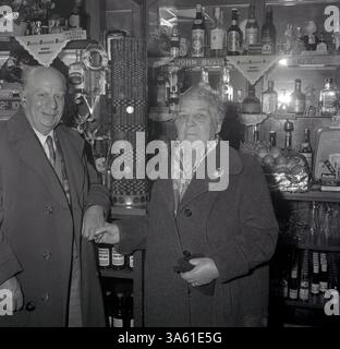 anni '1950, coppia storica, anziana nei loro cappotti in piedi in un pub bar vicino a una pila di monete,... una "torre delle monete", un modo divertente di raccogliere fondi di beneficenza spesso tenuti nei pub di quest'epoca. Una volta che il "penny pile" ha raggiunto una certa altezza, si sarebbe tenuta una cerimonia per abbatterlo, il denaro contato e dato a un ente di beneficenza locale o ospedale. Foto Stock