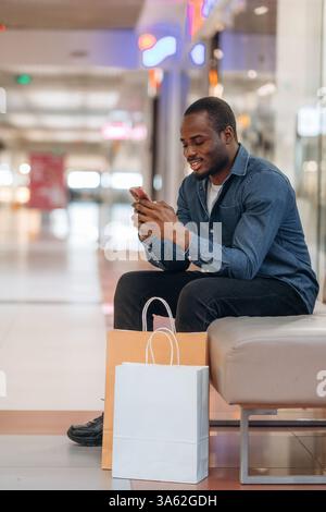 Vista laterale, seduto sulla panchina, tenendo in mano lo smartphone. L'uomo nero è nella sala del supermercato. Foto Stock