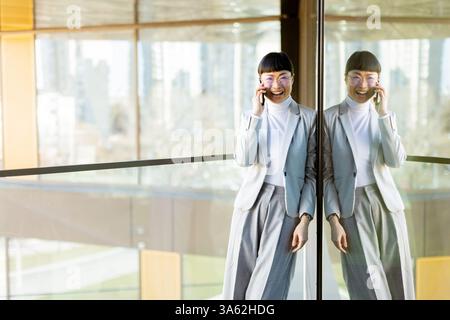 Una donna asiatica alla moda vestita con un abito sartoriale si staglia gioiosamente, chiacchierando al telefono mentre si appoggia a una parete di vetro. Foto Stock