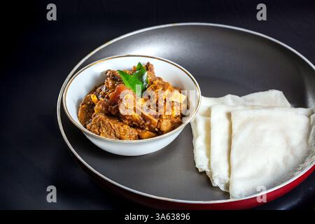 Pathiri di riso con masala di manzo o arrosto, una ricetta casalinga in stile Kerala, guarnita con foglie di curry e servita in una ciotola bianca contro uno sfondo nero Foto Stock