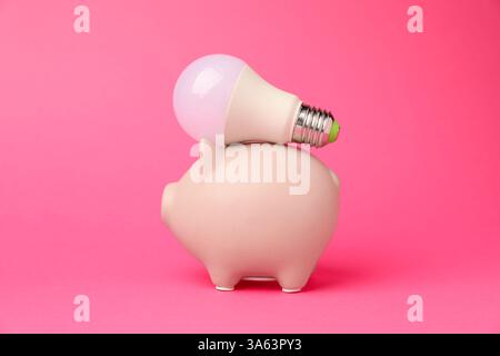 Salvadanaio con lampadina su sfondo rosa. Concetto di risparmio energetico Foto Stock