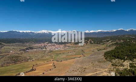 Campi coltivati e una piccola città sono annidati ai piedi di una maestosa catena montuosa innevata sotto un cielo blu vibrante Foto Stock