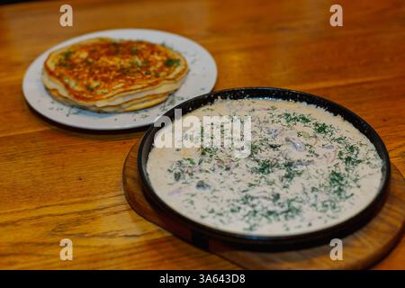 Frittelle fresche fritte su un piatto e sugo di carne in una padella con erbe e funghi panna acida Foto Stock