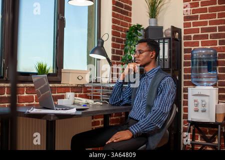 Giovane uomo d'affari seduto alla scrivania dell'ufficio con auricolari wireless, che guarda le presentazioni online sul suo personal computer. Un dipendente di sesso maschile di colore che lavora in un ambiente di lavoro a muro, frequenta corsi di formazione virtuali su notebook Foto Stock