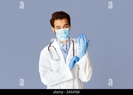 Protezione contro le malattie virali. Medico indiano che indossa maschera medica, camice da laboratorio e guanti di gomma su sfondo viola Foto Stock