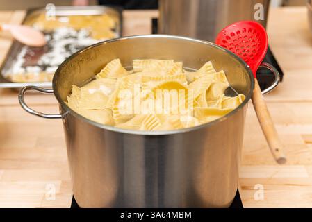 Una pentola in acciaio inox riempita con ravioli fatti in casa che cucinano in acqua bollente, con un cucchiaio rosso scanalato appoggiato sull'impugnatura. Foto Stock