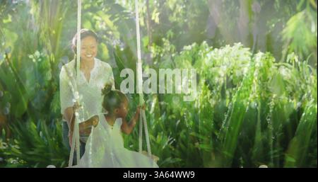 Spingendo la figlia sull'altalena in un giardino lussureggiante, la madre afroamericana sorride con gioia Foto Stock