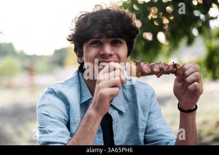 Giovane felice che tiene in mano uno spiedino di carne alla griglia all'aperto durante un barbecue estivo, sorridendo e gustando il cibo in un ambiente informale e sociale. Foto Stock