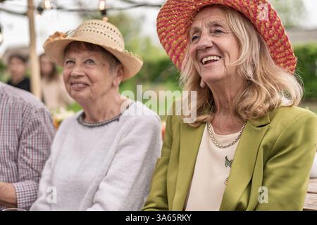Due allegre donne anziane che si godono una festa all'aperto indossando cappelli di paglia e sorridendo, catturate in un'atmosfera luminosa e rilassata. Foto Stock