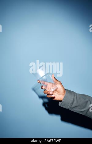 Mano con pelle chiara che tiene la tazza in plastica trasparente su sfondo azzurro creando ombre. Composizione semplice incentrata sul minimalismo e sull'oggetto monouso Foto Stock