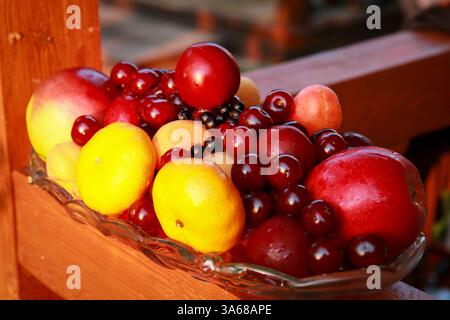 Una colorata disposizione di frutta fresca che include limoni, mele, ciliegie, prugne e albicocche disposte ordinatamente in un vaso di vetro su una terrazza di legno. Foto Stock