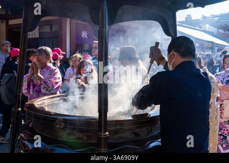 Uomo che accende incenso presso un bruciatore pubblico nel tempio Sensō-ji, Asakusa, Tokyo, circondato da visitatori. Foto Stock