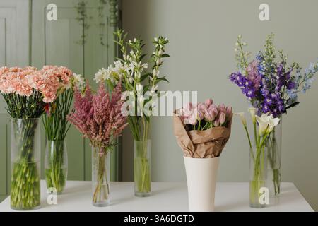 Elegante composizione floreale con fiori freschi e colorati in vasi di vetro su un tavolo bianco su un morbido sfondo verde, creando un'atmosfera serena, Foto Stock