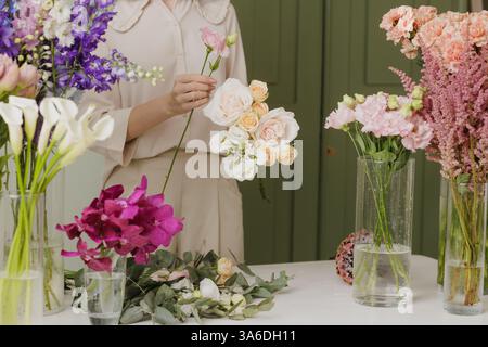 Fiorista donna che crea un delicato bouquet con fiori freschi in un laboratorio floreale, circondato da fiori colorati in vasi di vetro. negozio di fiori Foto Stock