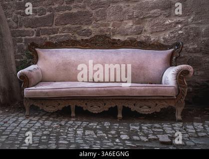 Un grande, vecchio divano di legno è seduto su una strada acciottolata. Il divano è coperto da una coperta bianca e viene abbandonato. La scena è di solitudine Foto Stock