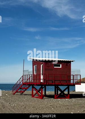 Una capanna rossa bagnino su palafitte si affaccia sull'oceano, circondata da sabbia e un cielo blu brillante con nuvole di neve. Foto Stock