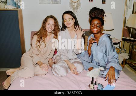 Ritratto di amici sorridenti e rilassanti in pigiama di seta e cotone seduti sul letto dopo una manicure a casa Foto Stock
