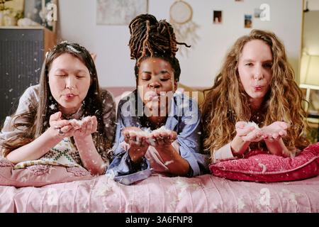 Il primo piano di tre adorabili giovani donne in pigiama sdraiate sul letto e che soffiano via piume di cuscini Foto Stock