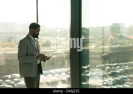 Un uomo ben vestito sorride mentre usa un tablet in un ufficio luminoso. La luce del sole proietta un caldo bagliore, mettendo in risalto la sua espressione focalizzata Foto Stock