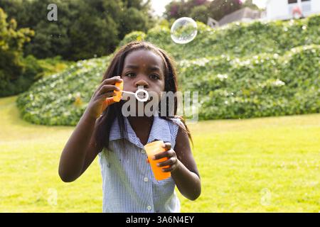 Ragazza afroamericana che soffia bolle nel parco, godendosi una giornata di sole all'aperto Foto Stock