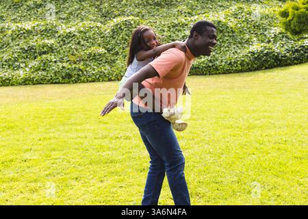 Padre che dà un giro in piggyback a figlia nel parco, entrambi ridendo gioiosamente Foto Stock