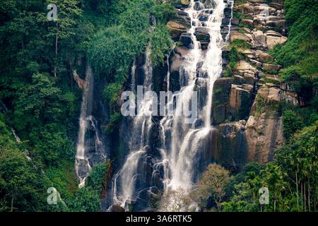 Una splendida cascata a più livelli scende lungo le scogliere rocciose di Ella, Sri Lanka, circondata da vegetazione lussureggiante e terreni agricoli terrazzati, creando un ambiente sereno Foto Stock