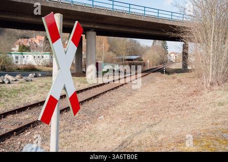 Attraversamento ferroviario contrassegnato da un cartello sotto un ponte autostradale. I binari si estendono in lontananza, circondati da alberi ed edifici. Simboleggia Foto Stock