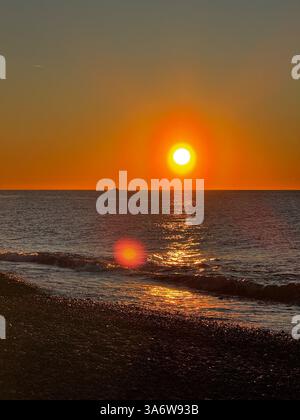 Un tramonto dorato sull'oceano, con una barca in lontananza e riflessi scintillanti sull'acqua e sulla spiaggia di ciottoli. Foto Stock