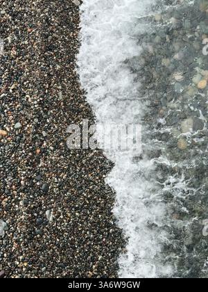 Vista aerea delle dolci onde oceaniche che scivolano su una spiaggia di ciottoli, mostrando contrasto tra acqua e pietre. Foto Stock