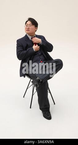 Foto a tutto corpo di un uomo d'affari asiatico seduto su una sedia e con una cravatta regolabile che sembra seduto su una sedia su sfondo bianco. Foto Stock