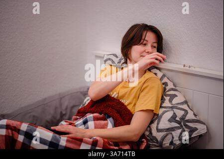 Una donna si siede a letto, avvolta da una coperta a quadri, e cerca con calma i suoi occhiali su uno scaffale vicino. Sembra rilassata e a suo agio in una camera da letto accogliente Foto Stock